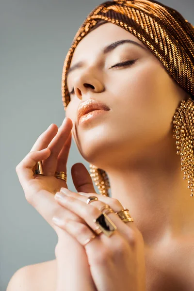 Mujer joven con los ojos cerrados, maquillaje brillante, anillos de oro y pendientes en turbante aislado en gris - foto de stock
