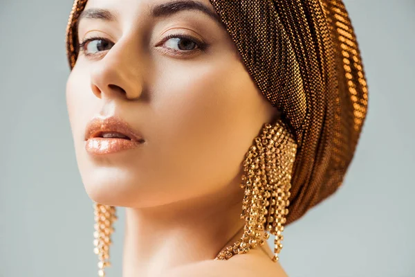 Junge schöne Frau mit glänzendem Make-up, Turban und goldenen Ohrringen, die in die Kamera schaut, isoliert auf grau — Stockfoto