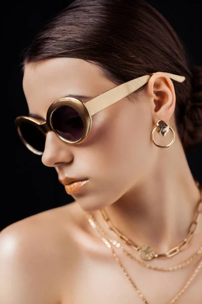 Mujer desnuda joven en gafas de sol, pendientes de oro y collares aislados en negro - foto de stock