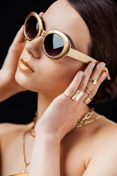 Jeune femme en lunettes de soleil, bagues dorées et colliers touchant les oreilles isolées sur noir — Photo de stock