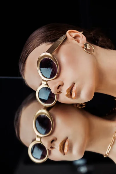 Mujer joven en gafas de sol, joyas de oro acostado en el espejo aislado en negro - foto de stock