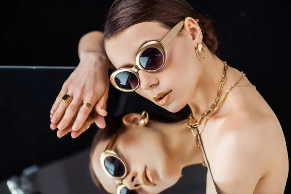 Mujer desnuda joven en gafas de sol, joyas de oro acostado en el espejo aislado en negro - foto de stock