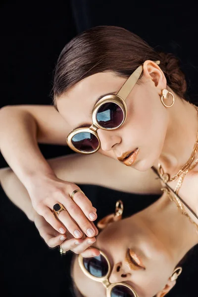 Young nude woman in sunglasses, golden jewelry lying on mirror isolated on black — Stock Photo