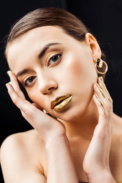 Mujer desnuda joven con maquillaje dorado y pintura dorada en los dedos tocando la cara aislada en negro - foto de stock