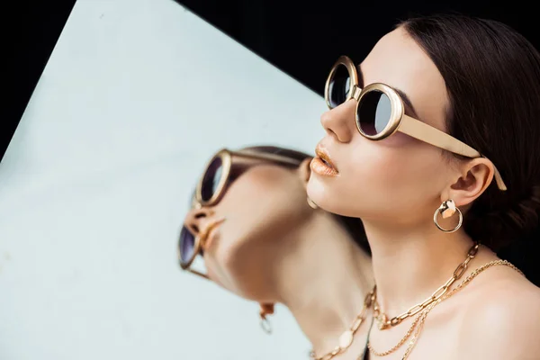 Young naked woman in sunglasses, golden necklaces holding mirror isolated on black — Stock Photo