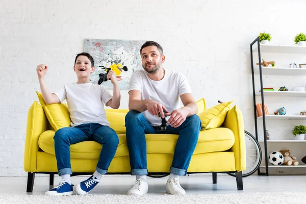 Animado filho torcendo enquanto joga vídeo game com o pai no sofá em casa — Fotografia de Stock