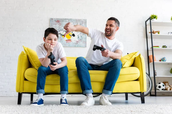 Padre mostrando el pulgar hacia abajo mientras está sentado con su hijo y jugando Videojuego en casa - foto de stock