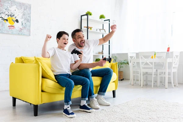 Emocionado padre e hijo vitoreando mientras juega videojuego en el sofá en casa - foto de stock