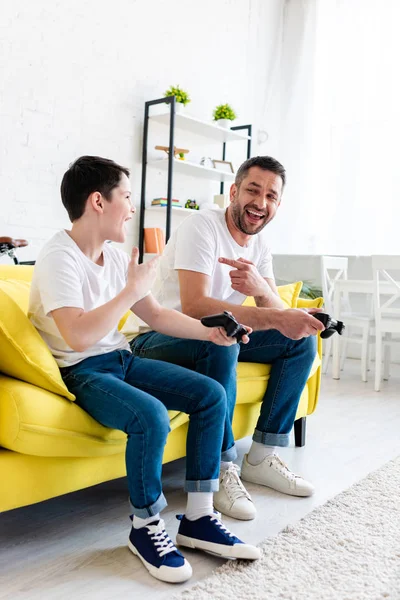 Pai animado e filho jogando Video Game no sofá e apontando com os dedos na sala de estar — Fotografia de Stock