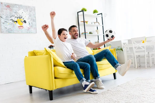 Animado pai e filho torcendo e assistindo jogo de esportes no sofá na sala de estar — Fotografia de Stock