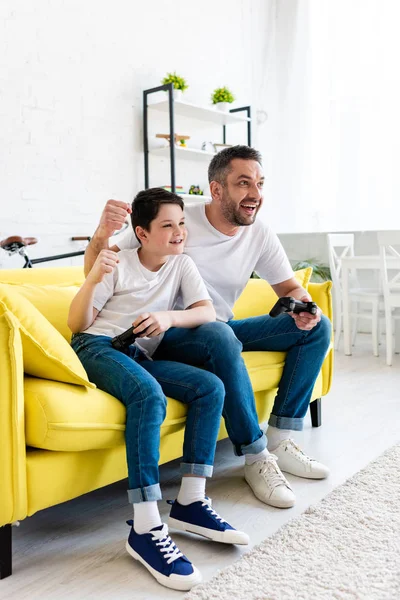 Padre e hijo emocionados animando mientras juegan videojuegos en el sofá en casa en la sala de estar - foto de stock