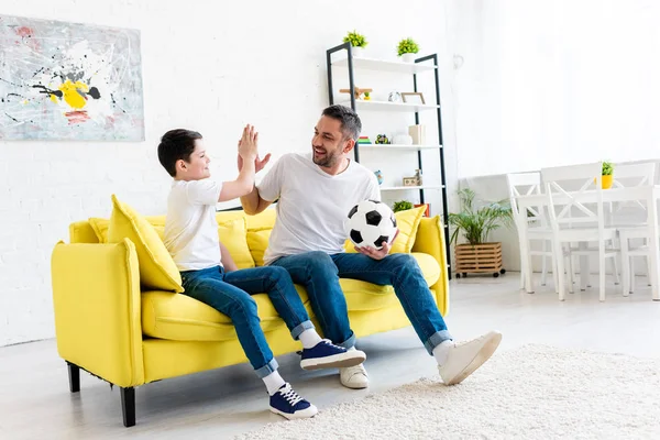 Батько і син роблять високі п'ять знаків, сидячи на дивані з футбольним м'ячем вдома — стокове фото