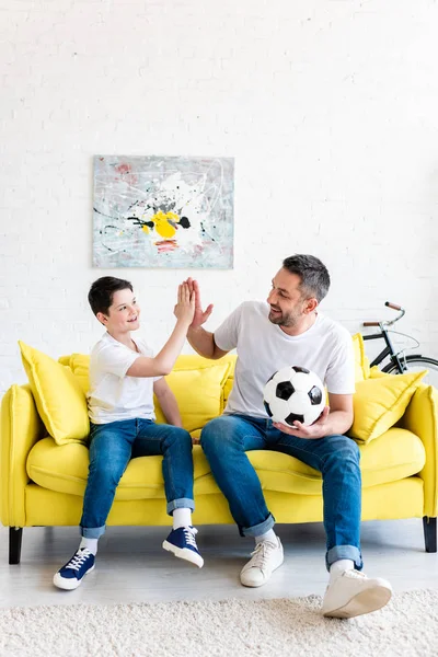 Padre e hijo haciendo alta cinco signo mientras está sentado en el sofá con pelota de fútbol en casa - foto de stock