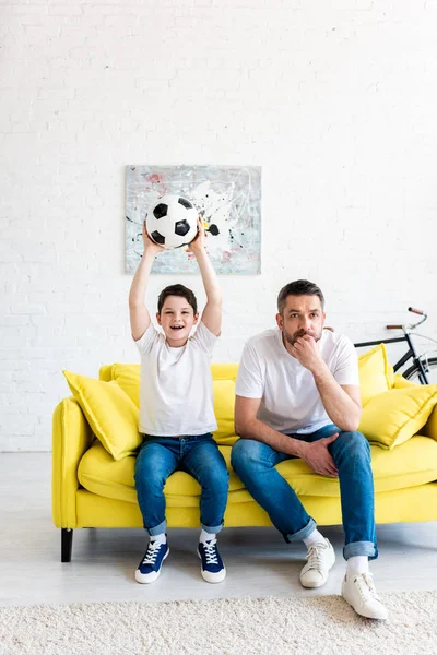 Hijo emocionado con pelota de fútbol viendo deportes partido con el padre en la sala de estar - foto de stock