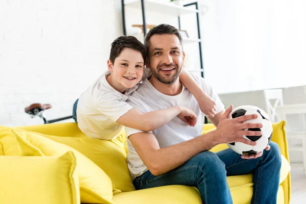 Feliz padre e hijo en el sofá con pelota de fútbol mirando a la cámara en casa - foto de stock