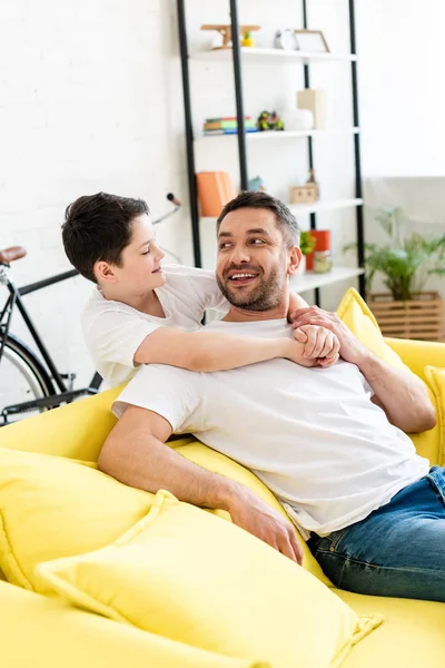 Filho abraçando sorrindo pai sentado no sofá em casa — Fotografia de Stock