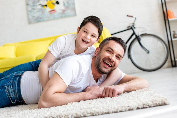 Щасливий батько і син лежать на килимі, дивлячись на камеру і обіймаючись вдома — стокове фото
