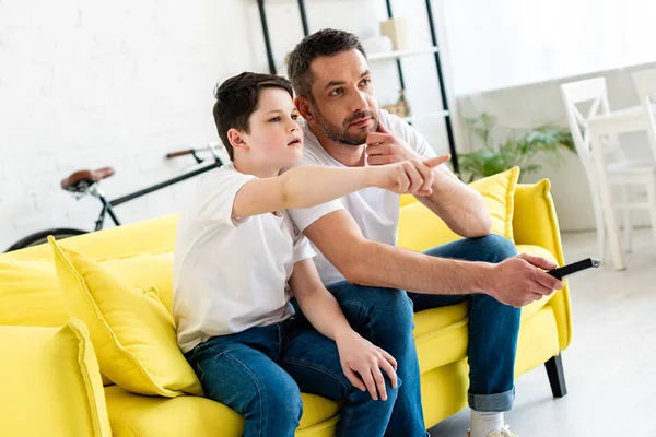Son pointing with finger while sitting on couch and watching tv with father at home — Stock Photo
