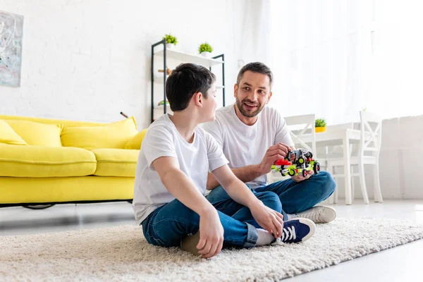 Feliz padre e hijo sentado en la alfombra y jugando con el coche de juguete en casa - foto de stock