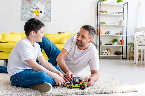 Улыбающиеся отец и сын сидят на ковре и играют с игрушечной машиной дома — стоковое фото