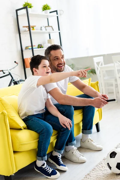 Hijo señalando con el dedo mientras está sentado en el sofá y viendo la televisión con el padre en casa - foto de stock