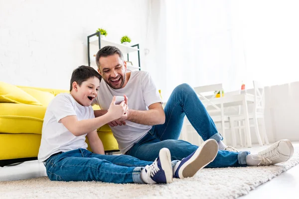 Padre e figlio eccitati seduti su tappeto e utilizzando lo smartphone in salotto — Foto stock