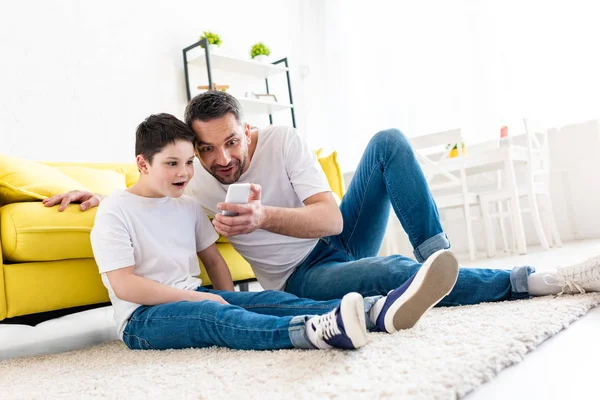 Sorprendido padre e hijo sentado en la alfombra y el uso de teléfono inteligente en la sala de estar - foto de stock
