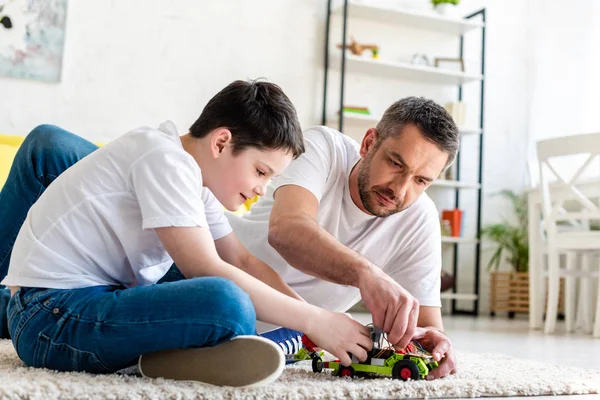 Padre e hijo sentados en la alfombra y jugando con coche de juguete en casa en la sala de estar - foto de stock