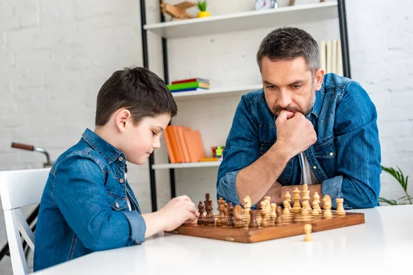 Padre e figlio pensieroso in denim che giocano a scacchi seduti a tavola a casa — Foto stock