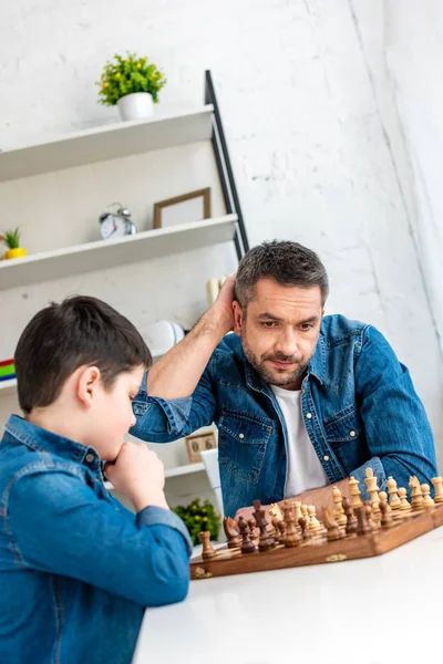 Padre e hijo guapos jugando ajedrez mientras están sentados en la mesa en casa - foto de stock