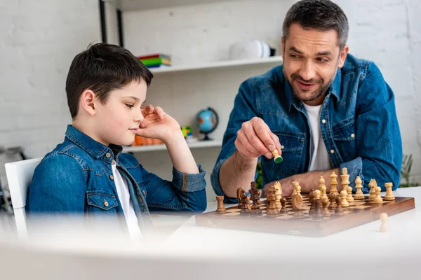 Enfoque selectivo de padre e hijo en el denim jugando ajedrez mientras está sentado en la mesa en casa - foto de stock