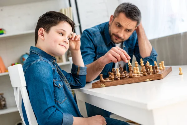 Vater und Sohn in Jeans spielen Schach, während sie zu Hause am Tisch sitzen — Stockfoto