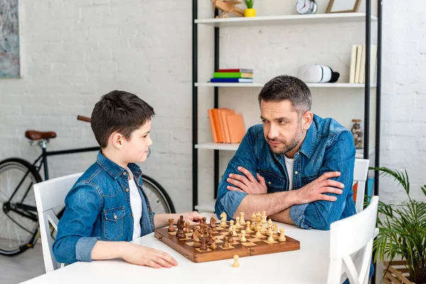 Vater und Sohn in Jeans spielen Schach, während sie zu Hause am Tisch sitzen — Stockfoto