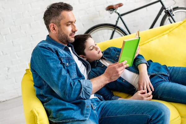 Pai e filho em jeans livro de leitura no sofá em casa — Fotografia de Stock