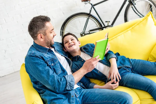 Happy father and son in denim reading book on couch at home — Stock Photo