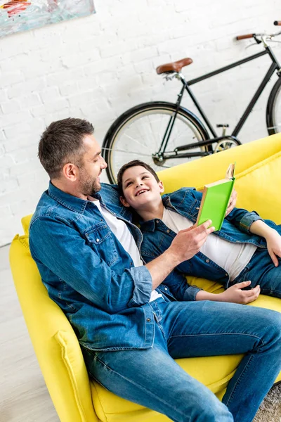 Happy father and son in denim reading book on couch at home — Stock Photo