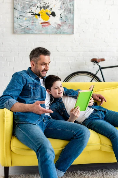 Улыбающиеся отец и сын в джинсах чтение книги на диване — стоковое фото