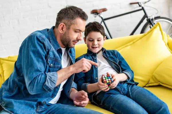 Père assis sur le canapé avec son fils et pointant du doigt le cube jouet — Photo de stock