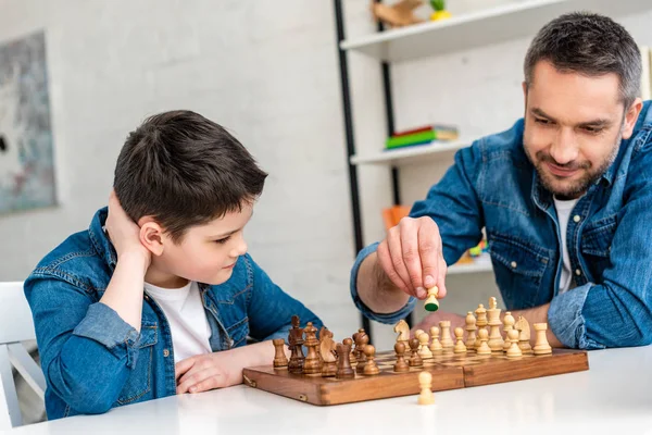 Père et fils en denim assis à table et jouant aux échecs à la maison — Photo de stock