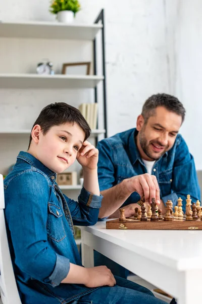 Pai e filho em ganga sentado à mesa e jogando xadrez em casa — Fotografia de Stock