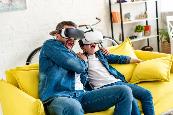 Отец и сын в наушниках VR кричат, испытывая виртуальную реальность на диване дома — стоковое фото