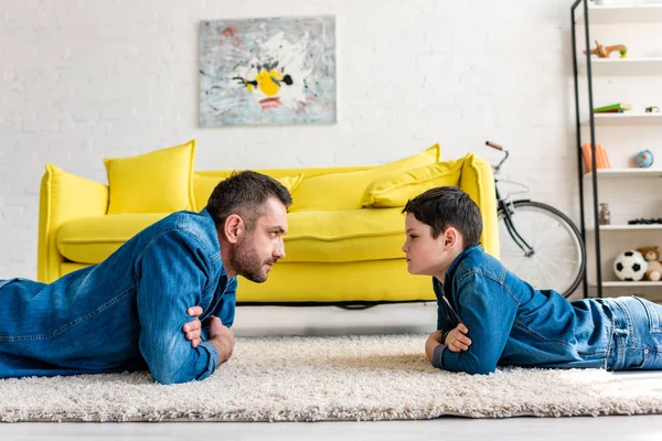 Father and son in denim with crossed arms lying on carpet and looking at each other at home — Stock Photo