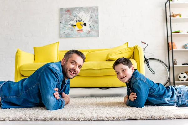 Feliz padre e hijo en denim con los brazos cruzados acostados en la alfombra y mirando a la cámara en casa - foto de stock