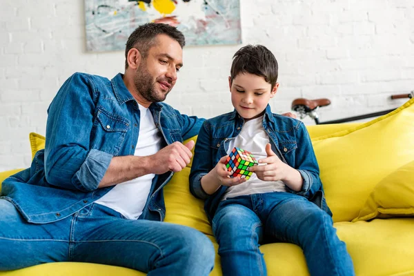 Padre e hijo sentados en el sofá y jugando con cubo de juguete en casa - foto de stock