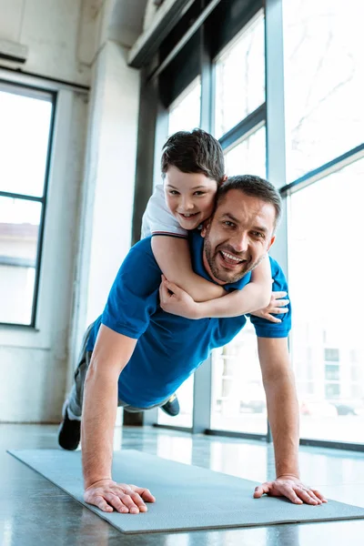 Feliz padre haciendo push up ejercicio con hijo en la espalda en el gimnasio - foto de stock