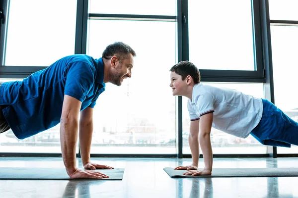 Pai e filho olhando um para o outro, enquanto fazendo push up exercício no ginásio — Fotografia de Stock