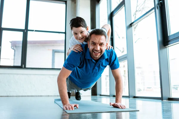 Щасливий батько робить Push Up вправи з сином на спині в тренажерному залі — стокове фото