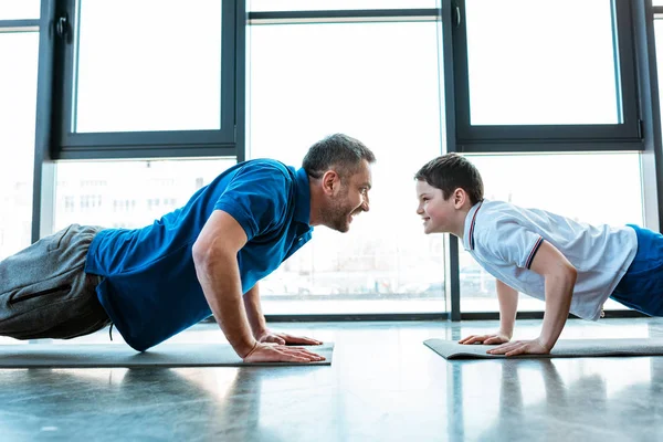 Padre e hijo mirándose mientras hacen ejercicio de push up en el gimnasio - foto de stock