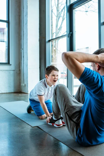 Hijo gritando mientras ayudar a padre haciendo ejercicio sentado en el gimnasio - foto de stock