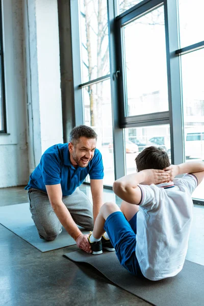 Father helping son sitting on fitness mat and doing sit up exercise at gym — Stock Photo